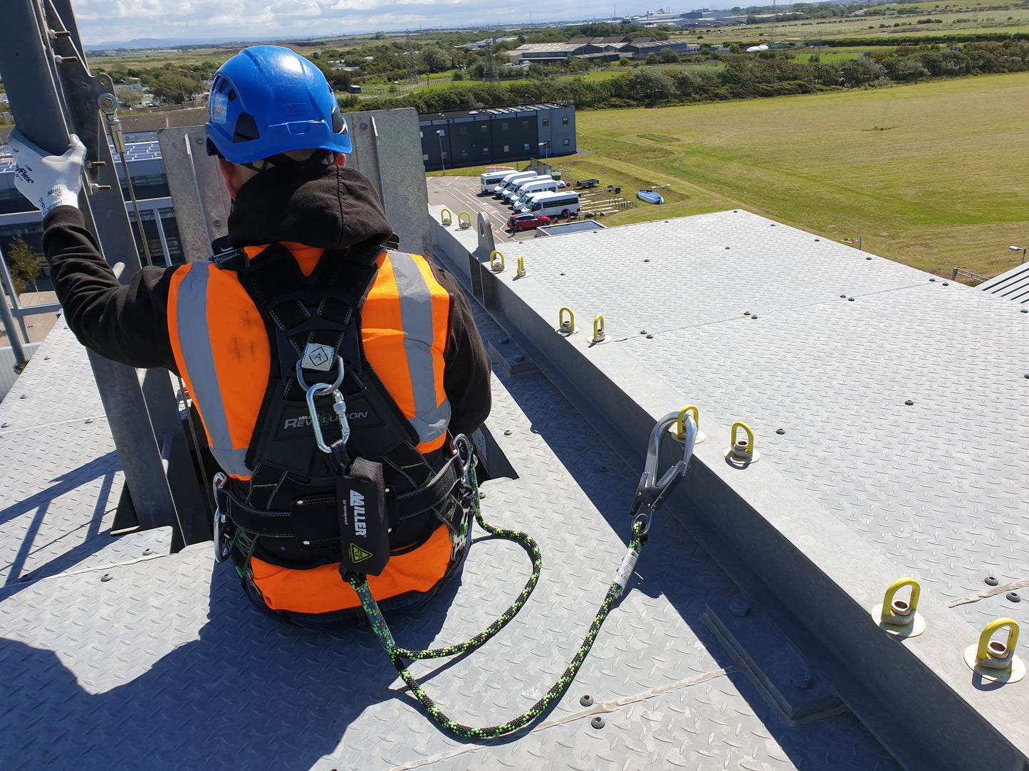 Person on a rooftop wearing safety harness attached to Honeywell Soll Turn Eye anchor point  by a rope lanyard with a view of vehicles and landscape in the background 